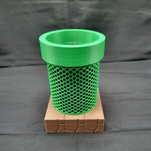 May include: A green 3D printed pipe with a mesh design and a brown base. The pipe is a popular video game item.