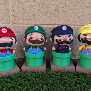 May include: Four handmade crochet figures of video game characters, each with a distinct hat and outfit. The figures are in green pipes, with brown bases. The characters include Mario, Luigi, Wario, and Waluigi.