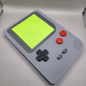 May include: A gray handheld video game console with a green screen, red buttons, and a black directional pad.