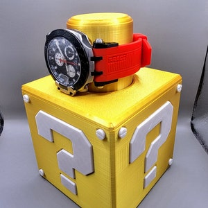 Puede incluir: Un bloque de interrogación amarillo impreso en 3D con un reloj de pulsera rojo y negro en la parte superior. El bloque tiene signos de interrogación blancos en cada lado.