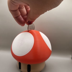 May include: A red and white mushroom-shaped piggy bank with a coin slot on top. A hand is placing a coin into the slot.