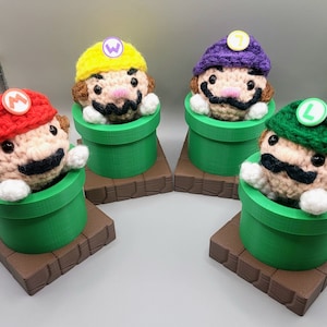May include: Four crocheted Mario characters, each wearing a different colored hat with a letter on it, are sitting in green pipes on brown bases. The characters are Mario, Luigi, Wario, and Waluigi.