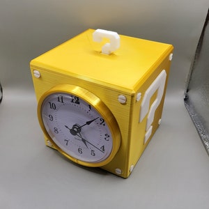 May include: A yellow cube-shaped clock with a white handle and a white question mark on the side. The clock face is white with black numbers and hands. The clock is a novelty item.