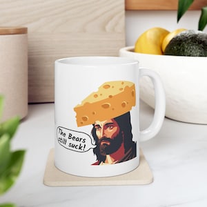 May include: White ceramic mug featuring a cartoon illustration of a man with a cheese wedge on his head. The man has a beard and long hair. A speech bubble says, "The Bears still suck!"