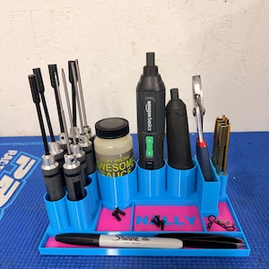 May include: A blue and pink tool organizer with various tools, including screwdrivers, a power tool, pliers, and a Sharpie marker. The organizer has the name "Nelly" printed on it and a bottle of "Scuff Racing Awesome Sauce".