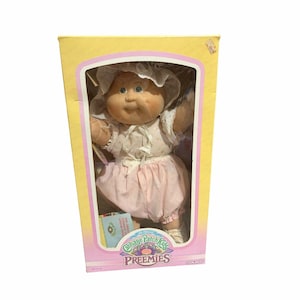 Peut inclure: Une poupée Cabbage Patch Kids Preemies dans son emballage d'origine jaune et rose. La poupée a un visage en vinyle souple, des yeux bleus et porte un bonnet blanc avec une bordure en dentelle, un haut à motifs assorti et un bloomer rose pâle.