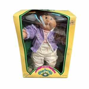 Könnte beinhalten: Eine Cabbage Patch Kids Puppe mit braunen Wollhaaren und gebräuntem Teint, bekleidet mit einer hellvioletten Reißverschlussjacke mit Rüschenbesatz über einem rosa Oberteil und einer weißen Hose. Die Puppe befindet sich in einem gelben Karton.