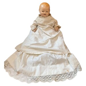 Puede incluir: Una muñeca de bebé de porcelana vintage que lleva un vestido blanco con ribete de encaje. La muñeca tiene una cara pintada con ojos azules y una tez rosada.