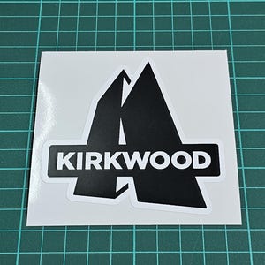 May include: A white sticker with a black graphic of two mountain peaks and the word "KIRKWOOD" in a white rectangular box. The sticker is on a green cutting mat.