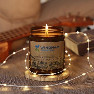 Könnte beinhalten: Eine brennende Sojakerze in einem braunen Glas mit einem Holzuntersetzer. Auf dem Etikett steht "Windham Mountain" und "Premium Scented Soy Candle Bring the Mountain Home". Die Kerze ist von einer Lichterkette umgeben.