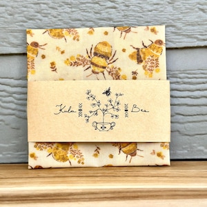 May include: A folded package featuring a bee and floral pattern. The package is wrapped with a band of brown paper with the text "Kula Bee" and a botanical illustration. The background is a light wood surface.