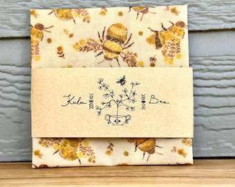May include: A folded package featuring a bee and floral pattern. The package is wrapped with a band of brown paper with the text "Kula Bee" and a botanical illustration. The background is a light wood surface.
