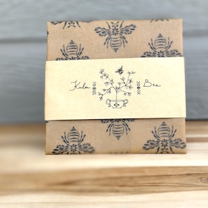 May include: A wrapped package with a tan background and a repeating pattern of navy blue bees. A cream-colored band with the words "Kula Bee" and a bee graphic wraps around the package. The package is sitting on a wooden surface.