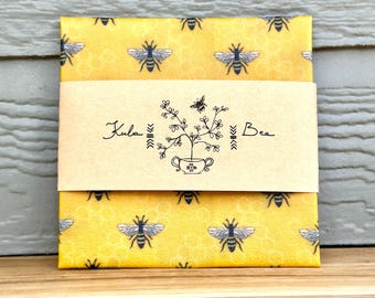 May include: A wrapped package featuring a yellow background with a honeycomb pattern and repeating bee illustrations. A brown paper band with the text "Kula Bee" and a bee-themed graphic wraps around the package. The package is sitting on a wooden surface.