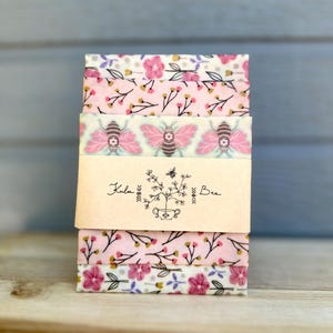 May include: A stack of reusable food wraps with floral and bee patterns. The wraps are pink, white, and purple, and are held together by a paper band with the text "Kula Bee" and a floral illustration. The wraps are made of a soft, flexible material.