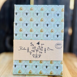 May include: A rectangular beeswax food wrap featuring a repeating pattern of bees and beehives on a light green background. A tan paper band wraps around the center, printed with the text Kula Bee, a floral illustration, and a small bee icon.