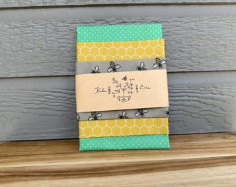May include: A stack of fabric squares in shades of yellow, green, and gray, featuring bee and honeycomb patterns. A brown paper band with the text "Kula Bee" wraps around the fabric. The fabric is stacked on a wooden surface.