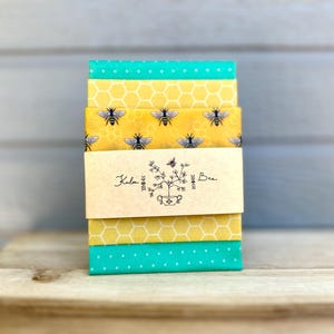 May include: A stack of fabric squares with bee and honeycomb patterns, and a teal polka dot pattern. A cream-colored label with the text "Kala Bee" and a floral illustration is wrapped around the fabric. The stack is on a wooden surface.