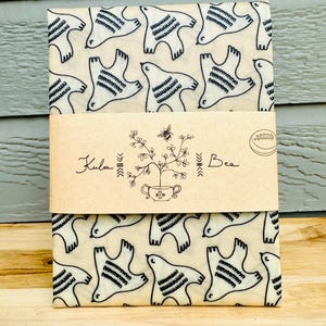 May include: A folded, rectangular package wrapped with a beige band. The package is covered in a repeating pattern of white birds with black details. The band features the text "Kula Bee" and a botanical illustration.
