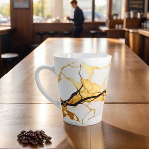 May include: White ceramic coffee mug with a gold and black marbled design. The mug is sitting on a wooden table with coffee beans scattered in front of it.