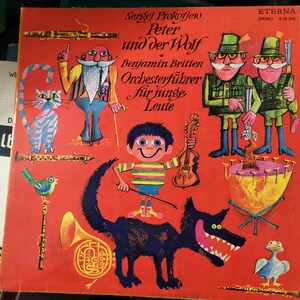 May include: A red, yellow, and blue illustrated record cover featuring a wolf, a cat, a bird, and a boy. The text on the cover reads "Sergej Prokofjew Peter und der Wolf Benjamin Britten Orchesterführer für junge Leute ETERNA STEREO 24.305".
