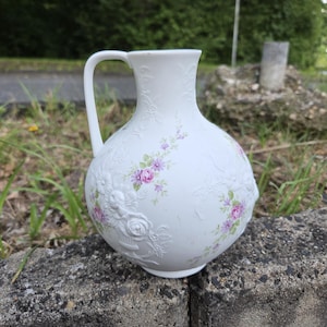 Puede incluir: Jarra de cerámica blanca con asa, adornada con diseños florales en relieve y delicados detalles de flores rosas y moradas. La jarra tiene un cuerpo redondeado y un cuello estrecho, ideal para servir bebidas.