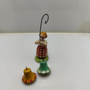 Könnte beinhalten: Eine Holzfigur einer Person, die einen Hirtenstab hält und ein rot-weiß kariertes Kleid trägt. Neben der Figur befinden sich zwei kleine glockenförmige Ornamente. Eines ist gelb mit roten Punkten, das andere grün mit weißen Punkten.