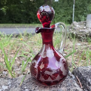 Könnte beinhalten: Eine rubinrote Glasdekanter mit einem Griff und einem Stopfen aus klarem Glas. Die Karaffe hat geätzte Designs von Laub und einem Hirsch. Der Stopfen hat eine runde Form mit geätzten Designs. Die Karaffe steht auf einem grauen Stein.