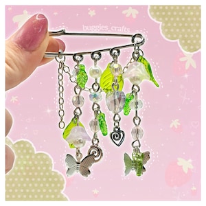Puede incluir: Un imperdible decorativo con colgantes. El imperdible tiene un acabado plateado y está adornado con hojas verdes, cuentas transparentes e iridiscentes, mariposas y un corazón. El texto "buggies_crafts" es visible en el imperdible.