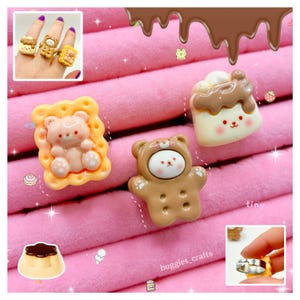 May include: Three rings featuring bear-shaped designs. One ring is a peach-coloured bear on a scalloped biscuit base, one is a brown bear wearing a hood, and one is a white bear topped with brown icing. The rings have adjustable metal bands.