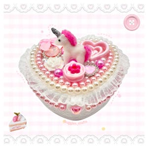 May include: A heart-shaped trinket box decorated with a white and pink unicorn figurine, pink and white pearls, a small pink bow, a white rose, and various heart-shaped charms. The lid is trimmed with white lace and features the text buggies_crafts.