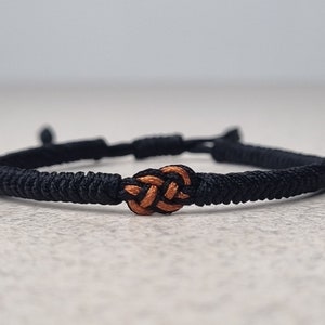 May include: Black woven bracelet with an orange knot in the center. The bracelet is adjustable and appears to be made of a durable, cord-like material. The knot is a focal point, adding a decorative element to the simple design. The bracelet is suitable for everyday wear.