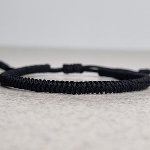 Op de afbeelding: Een zwarte gevlochten koord armband met een eenvoudige knoop sluiting.