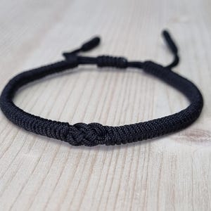 Op de afbeelding: Een zwarte gevlochten koord armband met een geknoopte sluiting.