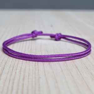 Peut inclure: Un bracelet violet vif avec un design de cordon à double brin. Le bracelet comporte des nœuds réglables pour un ajustement personnalisé. Le cordon est de couleur violet vif uni, présenté sur une surface en bois clair.