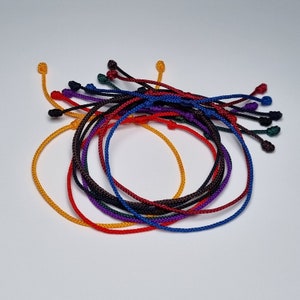 Può includere: Una collezione di braccialetti intrecciati colorati in rosso, arancione, giallo, verde, blu, viola e nero.