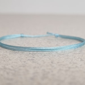 Pode incluir: Um bracelete de cordão trançado azul claro. O bracelete é feito de um cordão fino e macio e é amarrado com um nó simples.