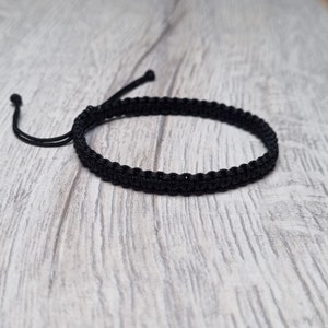 Könnte beinhalten: Ein schwarzes geflochtenes Armband mit verstellbarem Verschluss. Das Armband besteht aus einem dünnen, strapazierfähigen Band und ist etwa 18 cm lang.