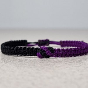 May include: A black and purple braided friendship bracelet with a knot in the center.