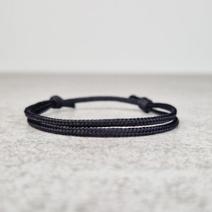 Peut inclure: Bracelet en corde noire avec une fine ligne blanche au centre. Le bracelet est noué aux deux extrémités pour l'ajuster. Design minimaliste, adapté au quotidien. Fabriqué avec un matériau durable.