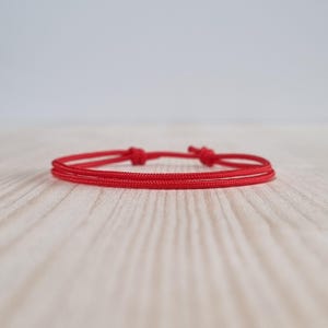 Può includere: Un braccialetto rosso minimalista con chiusura annodata. Il braccialetto è realizzato con un cordoncino sottile e intrecciato e poggia su una superficie di legno chiaro. Il design è semplice ed elegante, adatto all'uso quotidiano.