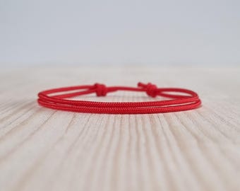 May include: A minimalist red bracelet with a knotted closure. The bracelet is made of a thin, woven cord and lies flat against a light-colored wooden surface. The design is simple and elegant, suitable for everyday wear.