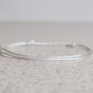 May include: A white cord bracelet with a simple design. The bracelet is made of two strands of cord that are tied together at the ends.