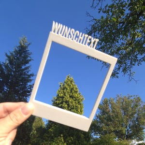Könnte beinhalten: Ein weißer, rechteckiger, 3D-gedruckter Rahmen in Form eines Sofortbildfotos, der am oberen Rand das Wort WUNSCHTEXT in Großbuchstaben eingeprägt hat. Der Rahmen hat eine glatte, matte Oberfläche und eine hohle Mitte zum Betrachten von Objekten.
