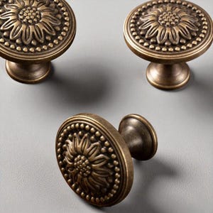 May include: Three antique bronze cabinet knobs. Each knob features a sunflower design in the center, surrounded by a beaded border. The knobs have a round shape and a short, cylindrical stem. The finish is a warm, aged bronze.