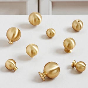 Puede incluir: Una colección de pomos dorados para armarios de varios tamaños. Los pomos tienen un diseño esférico con un acabado metálico liso. Algunos tienen una base cilíndrica, otros una base más decorativa. Los pomos están dispuestos sobre una superficie blanca.