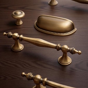 Puede incluir: Primer plano de herrajes de gabinete de latón antiguo, que incluyen un pomo, un tirador de taza y varios tiradores. El hardware tiene una estética vintage con detalles ornamentados y un tono dorado cálido. El hardware se muestra sobre una superficie de madera oscura.