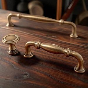 May include: Close-up of antique brass cabinet hardware. The set includes two curved handles and a round knob. The hardware has a vintage, aged appearance with a warm, golden-brown colour. The handles are approximately 15 cm long.