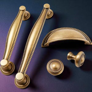 May include: A selection of antique brass cabinet hardware, featuring two long pull handles, a cup pull, a round knob, and a smaller knob. The hardware has a beaded edge design and a warm, aged finish, suitable for kitchen or furniture renovations.