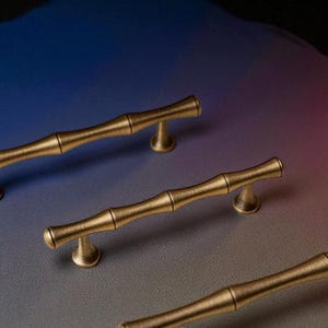 Op de afbeelding: Close-up van verschillende bronskleurige kastgrepen met een bamboe-ontwerp. De grepen zijn gerangschikt op een gradiëntachtergrond van blauw, paars en grijs. Elke greep heeft een cilindrische handgreep met een getextureerd oppervlak en is gemonteerd op twee kleine, afgeronde voetjes.